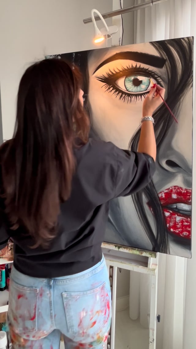 Didi Konings staat met haar rug naar de camera en schildert met een dun penseel het oog van een groot vrouwenportret.
